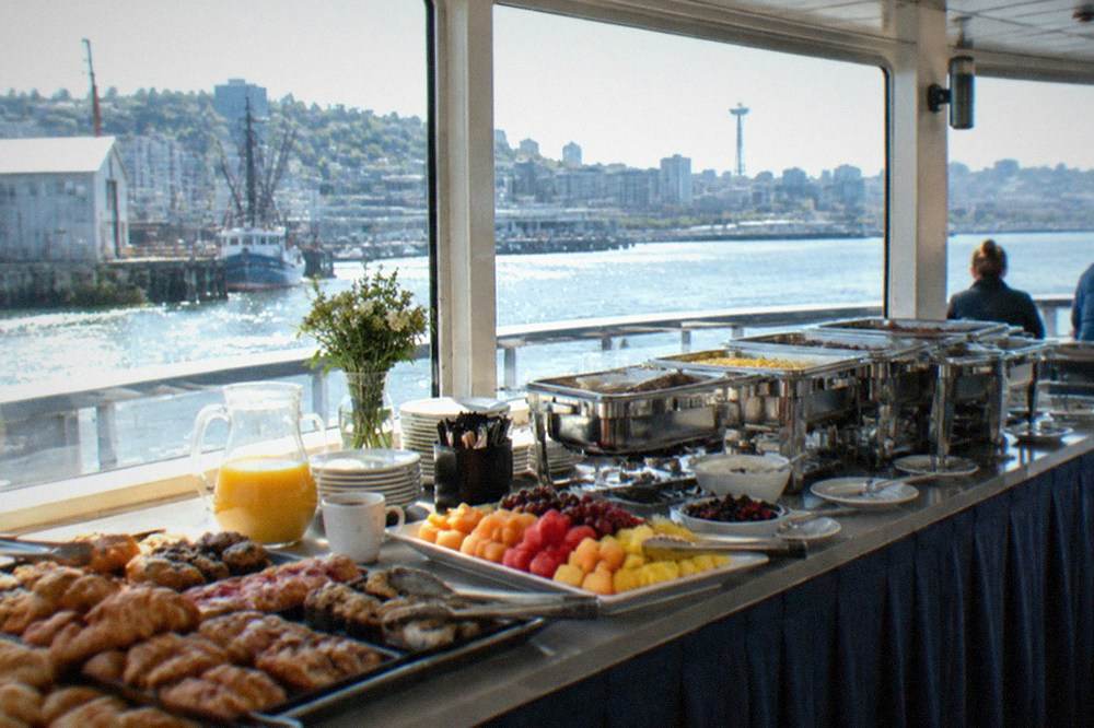 A brunch buffet table on a boat deck with serving trays of pastries and fruit, with lake water visible through the windows behind