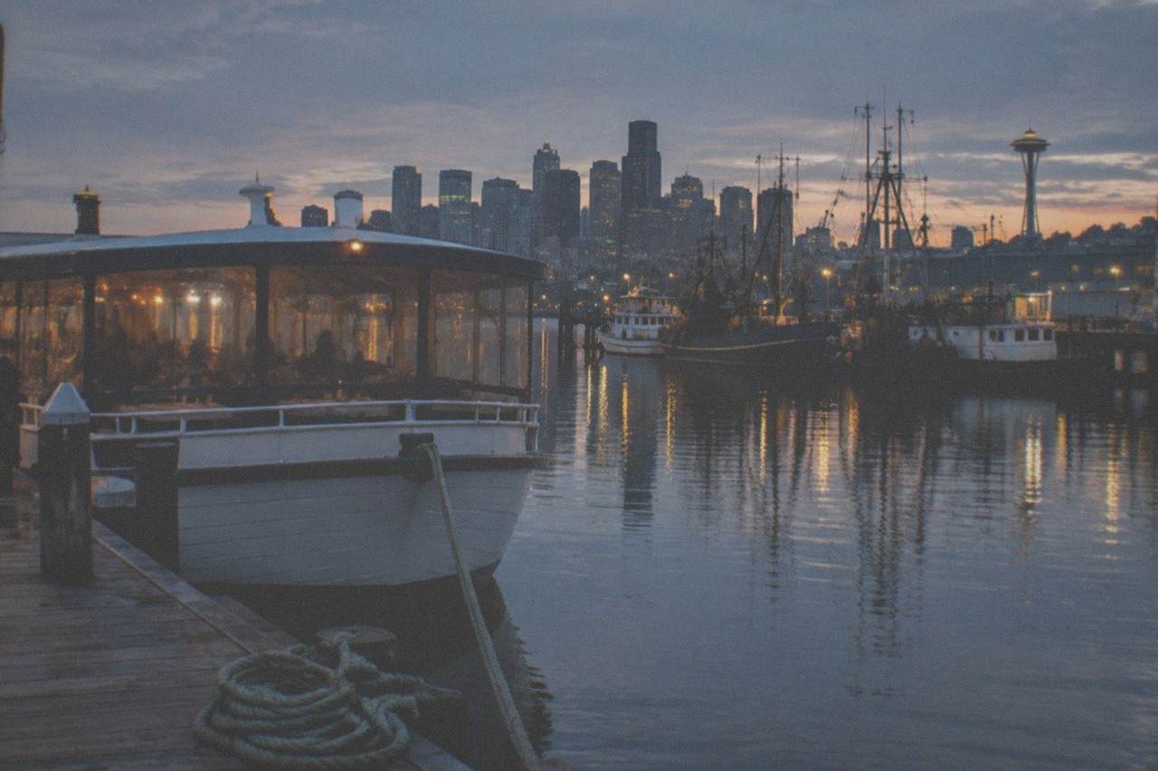 A covered dinner cruise boat moored at a Lake Union dock at dusk, with city lights reflected on the water