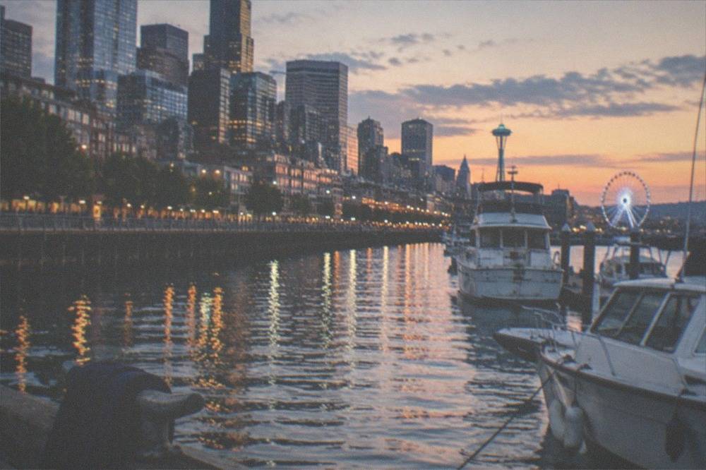 A waterfront pier in Seattle at dusk with city lights reflecting on the bay and moored boats in the foreground.