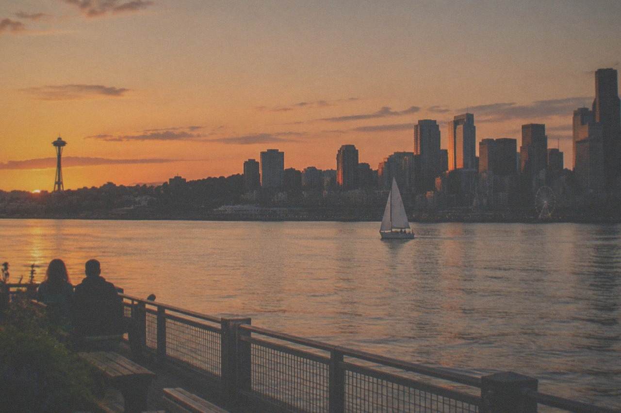 A sailboat on Elliott Bay at sunset with the Seattle skyline in silhouette behind calm water.