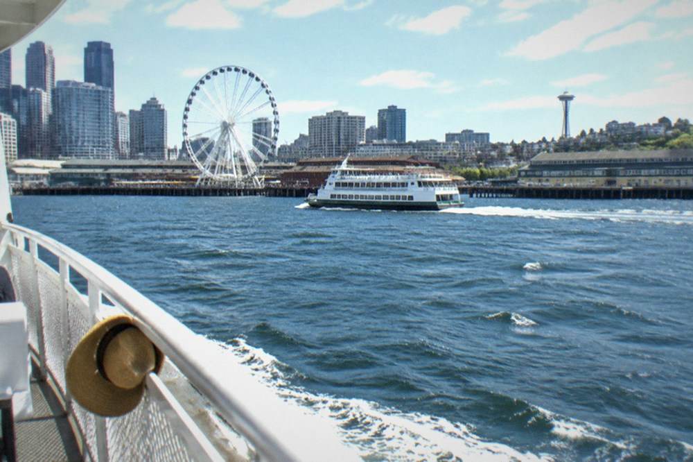 A tour boat passing the Seattle Great Wheel and waterfront piers on a bright afternoon on Elliott Bay.