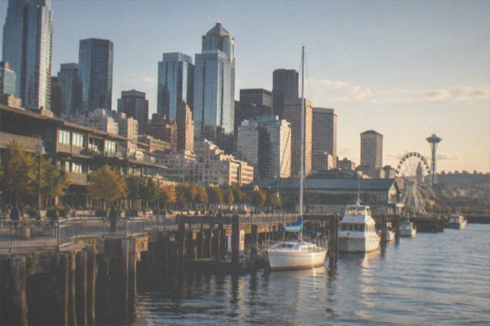 The central Seattle waterfront promenade and piers seen from the water, with the city skyline rising above the bay.