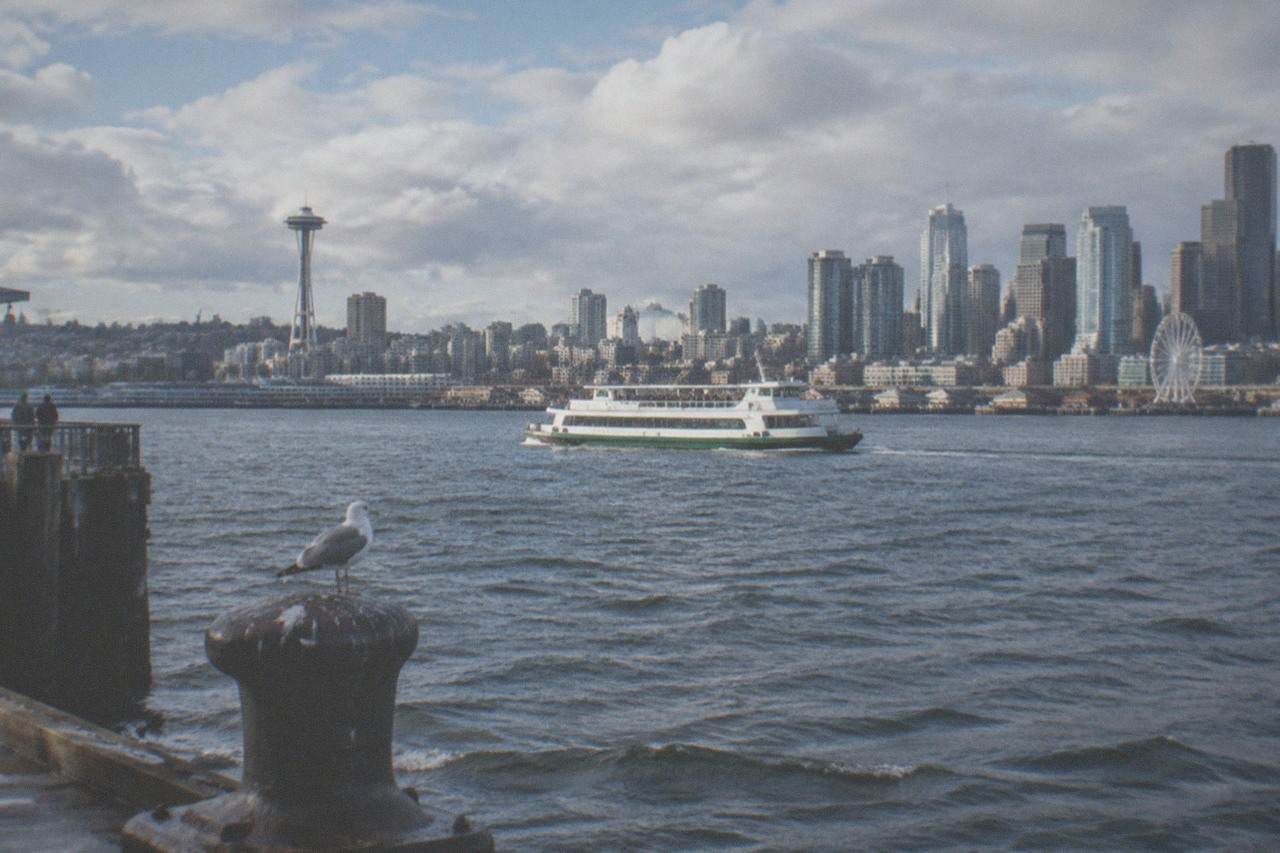A sightseeing boat crossing Elliott Bay with the Seattle skyline and Space Needle behind it under a partly cloudy sky.
