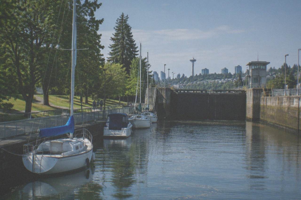 Sailboats and small motorboats tied along a tree-lined canal beside a large concrete lock gate