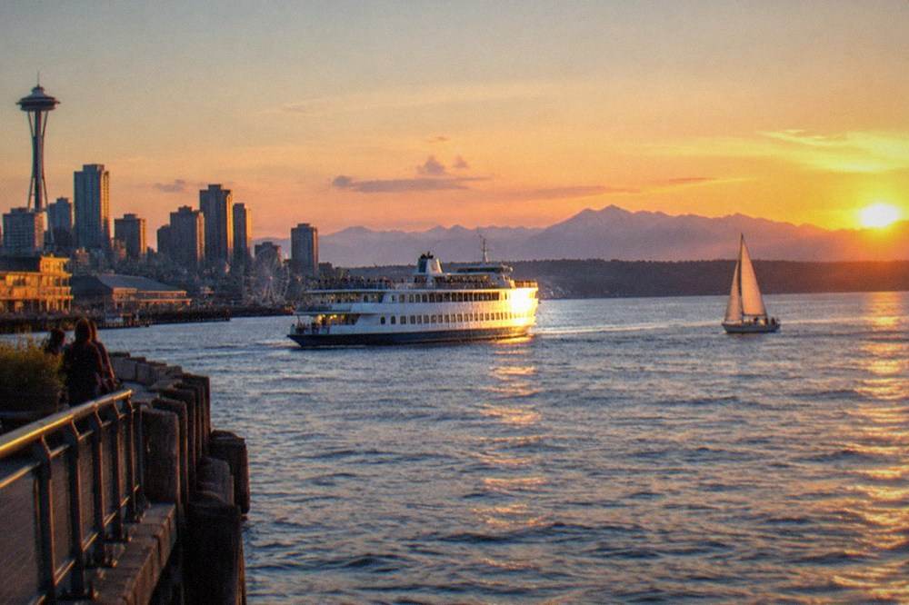 A tour boat and a sailboat on Elliott Bay under a summer sunset with the Olympic Mountains in the distance