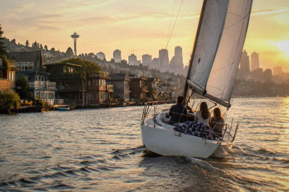 A sailboat heeling gently on Lake Union at golden hour, with houseboats and the Seattle skyline in the background