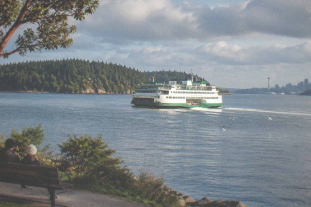 A ferry crossing a wide channel between forested islands under a partly cloudy sky