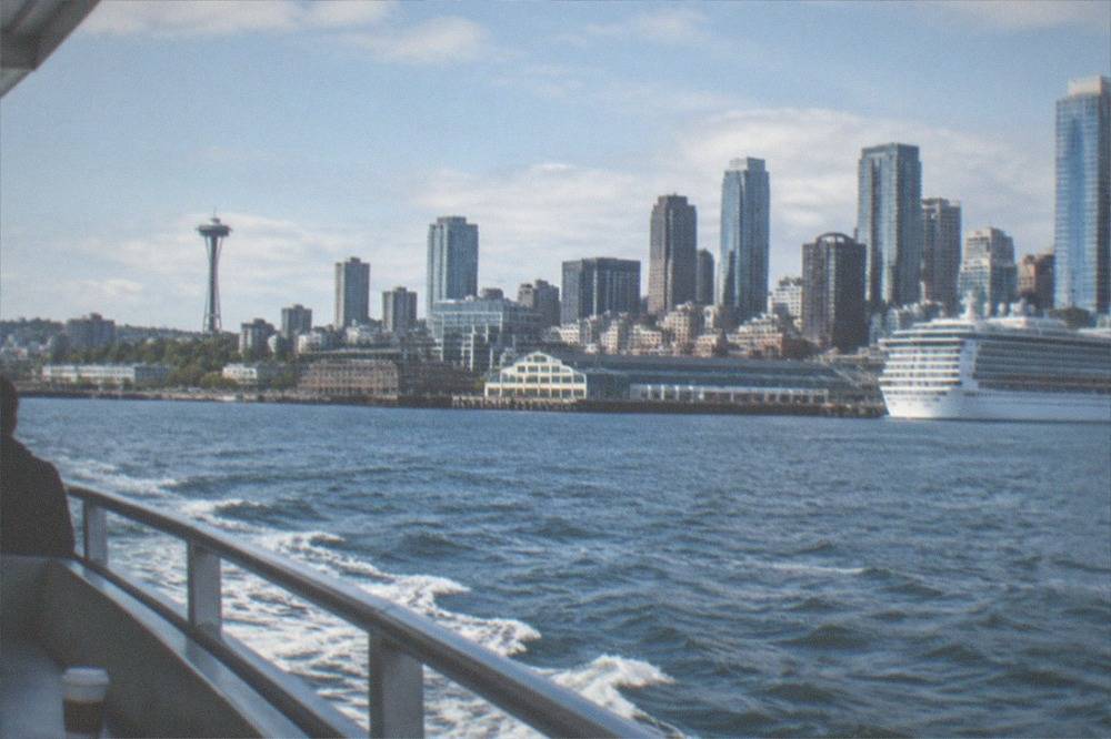 A small tour boat cruising on Elliott Bay with the Seattle skyline and a moored cruise ship in view.