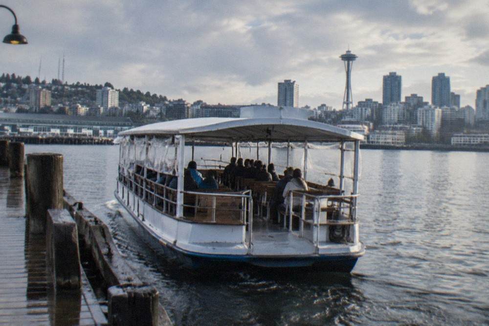 A covered sightseeing boat departing a downtown Seattle pier with passengers seated along both rails