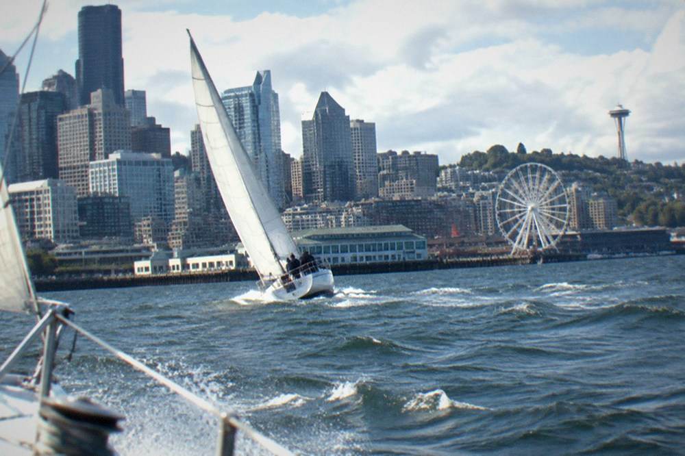 A sailboat heeling on Elliott Bay with the Seattle Great Wheel and downtown skyline in the background
