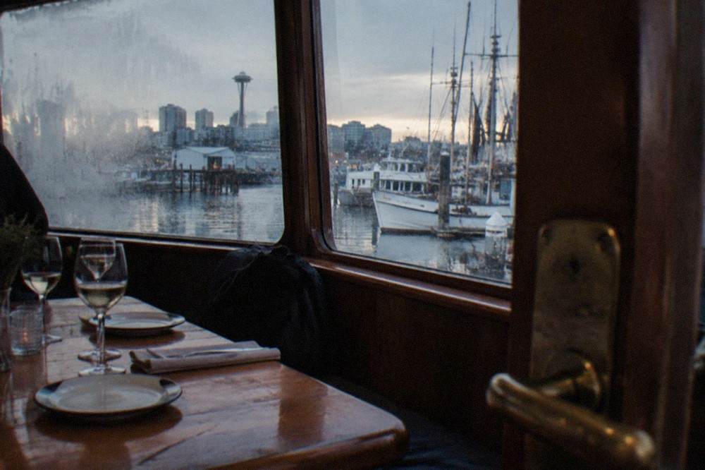 A dinner table set with glasses and plates inside a boat salon window at dusk