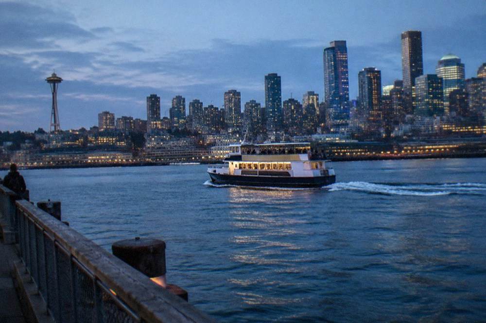 A small cruise boat crossing a city harbor at dusk with office towers behind it