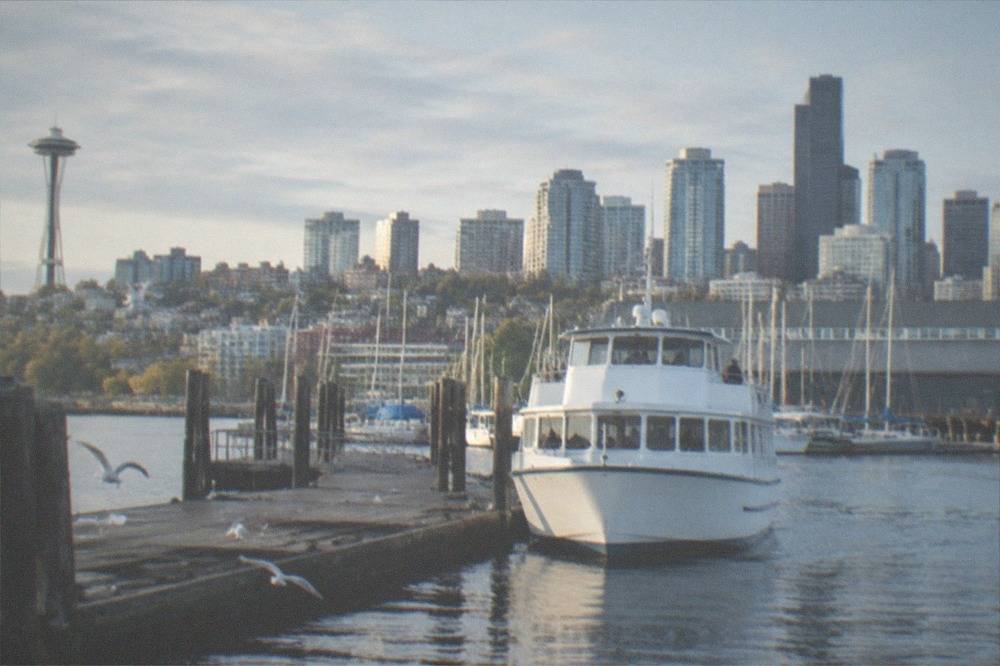 A tour boat departing a Seattle pier on a calm morning with moored masts and downtown buildings behind it
